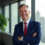 Portrait of a confident older businessman with gray hair wearing a blue suit and red tie, standing with arms crossed in a bright office